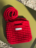 A bright red hand-crocheted shoulder bag with a matching crocheted strap, styled on a matte olive green surface with a white pebble background. A small white label on the front reads 'THIS DAY STUDIO'.