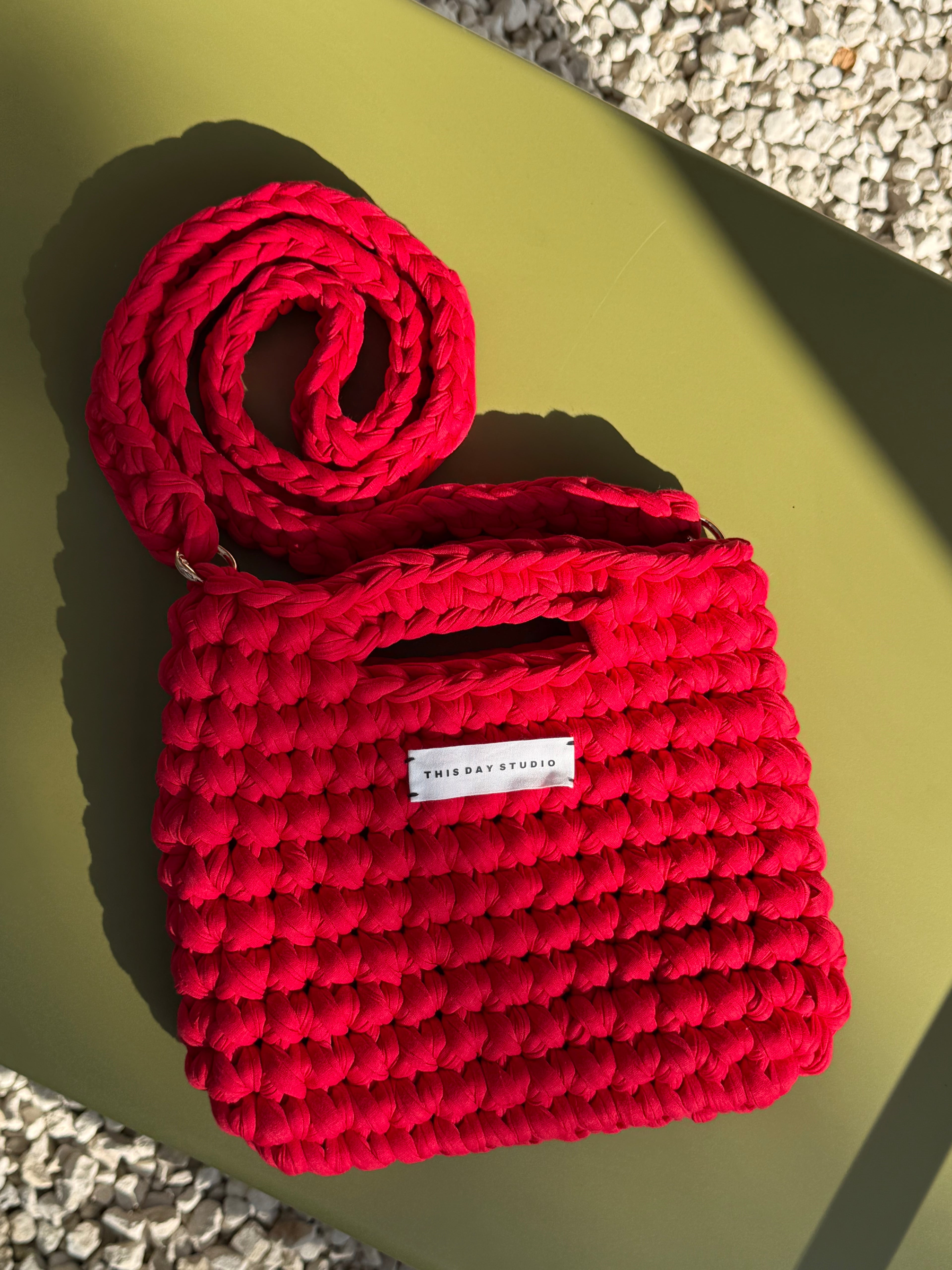 A bright red hand-crocheted shoulder bag with a matching crocheted strap, styled on a matte olive green surface with a white pebble background. A small white label on the front reads 'THIS DAY STUDIO'.