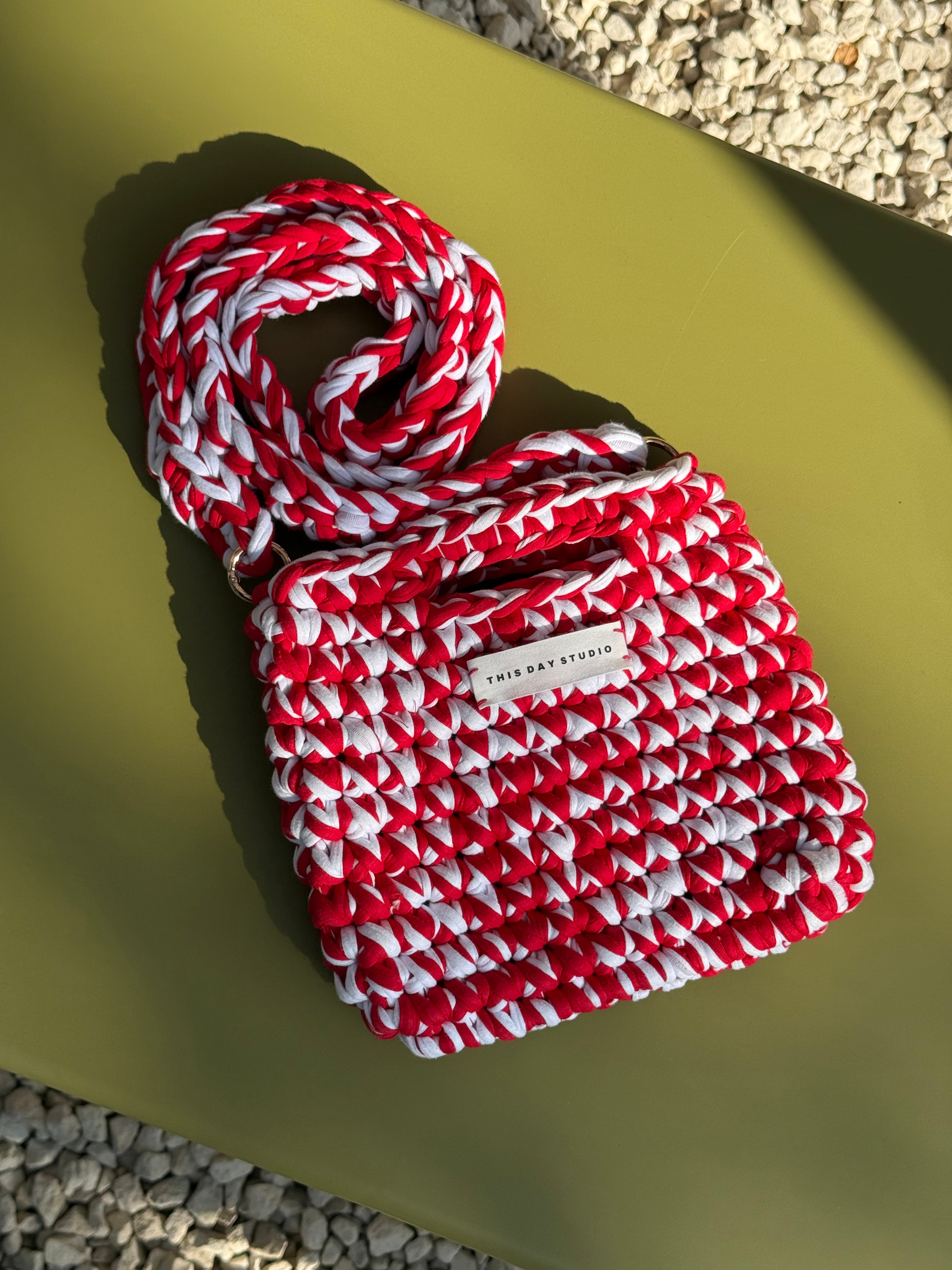 A woven crossbody bag with a thick braided strap in red and white, featuring a small white brand label that reads 'THE BAG STUDIO'. The bag is photographed from a top-down angle on a solid olive green surface with gravel visible in the corners.