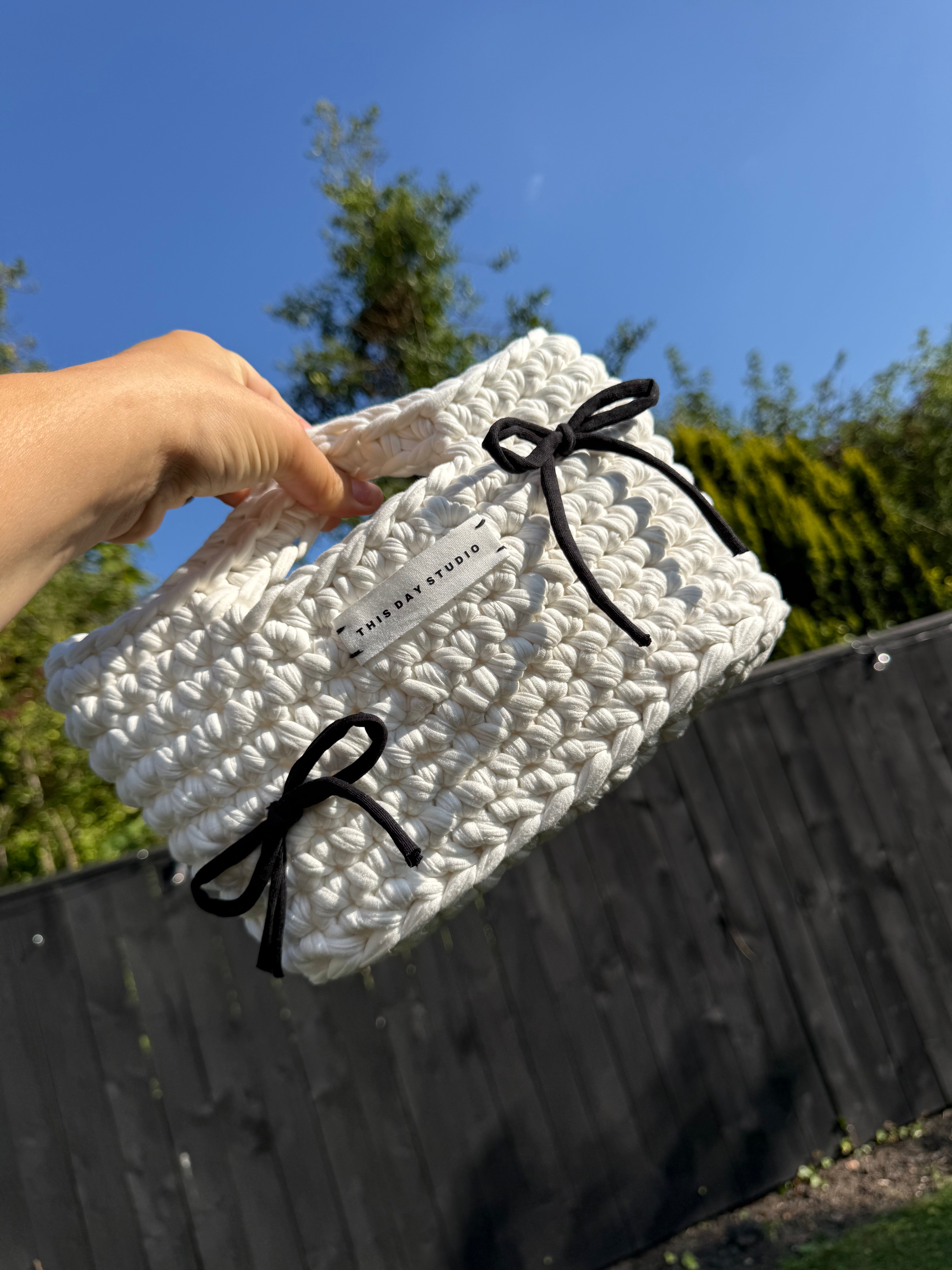 A hand holds a white chunky crocheted handbag with black ribbon bows against an outdoor background of a wooden fence, green trees, and blue sky. A brand tag reading 'THINLAAR STUDIOS' is visible on the bag.