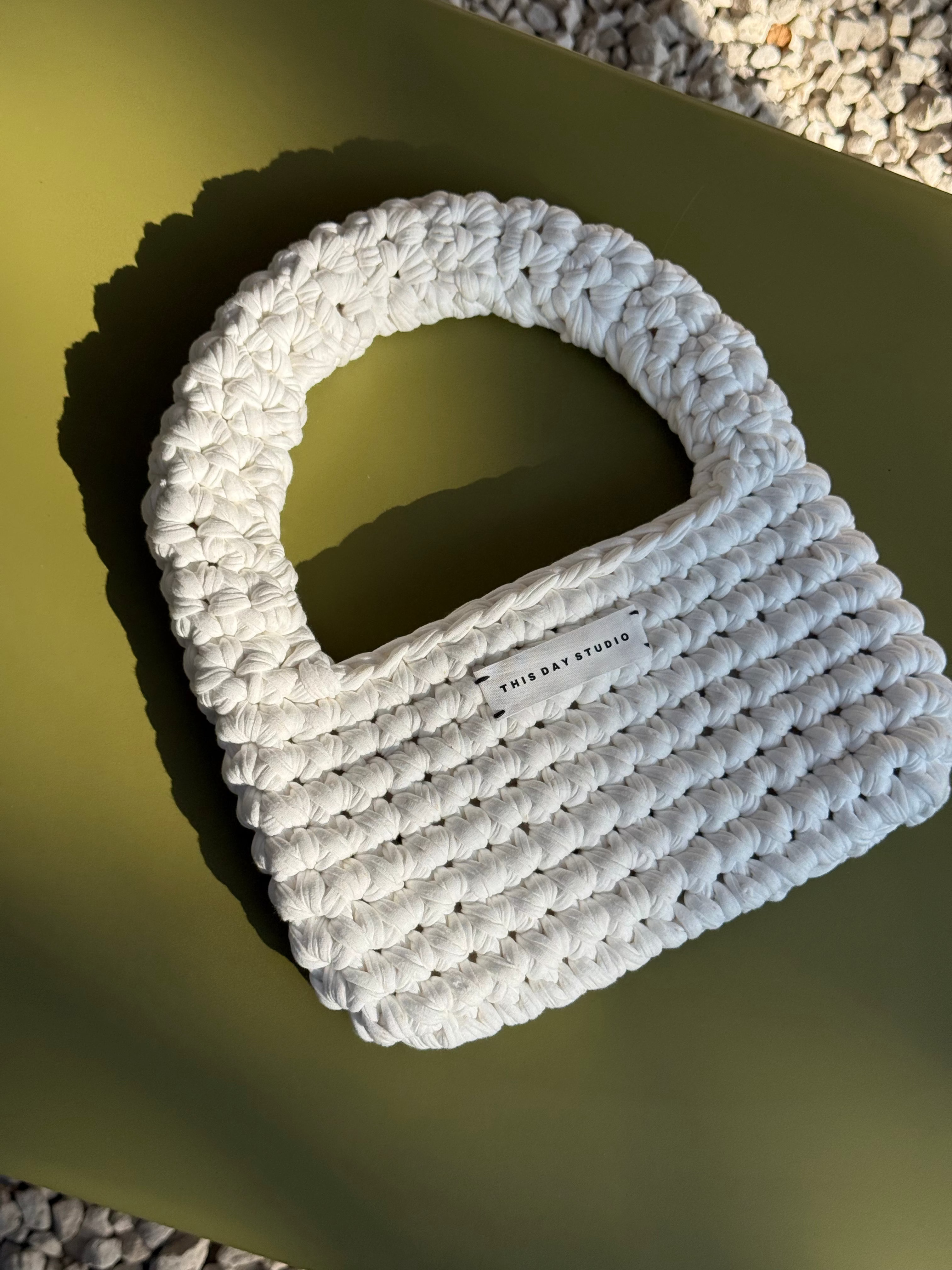 A chunky white crocheted shoulder bag with a large integrated handle, resting on a dark olive green surface with a shadow cast to the right.